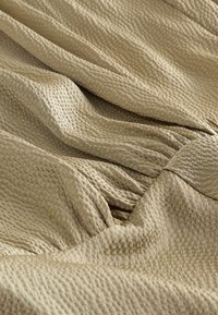 Beige gestructureerde stof met een geraffineerd, geplooid ontwerp en lichte plooien die zachte schaduwen en diepte creëren.