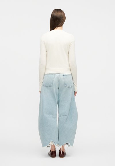 Άτομο με μακριά καστανά μαλλιά φοράει λευκό πουλόβερ, φαρδιά ανοιχτό μπλε τζιν με φθορές και καφέ παπούτσια, στέκεται μπροστά σε απλό φόντο.