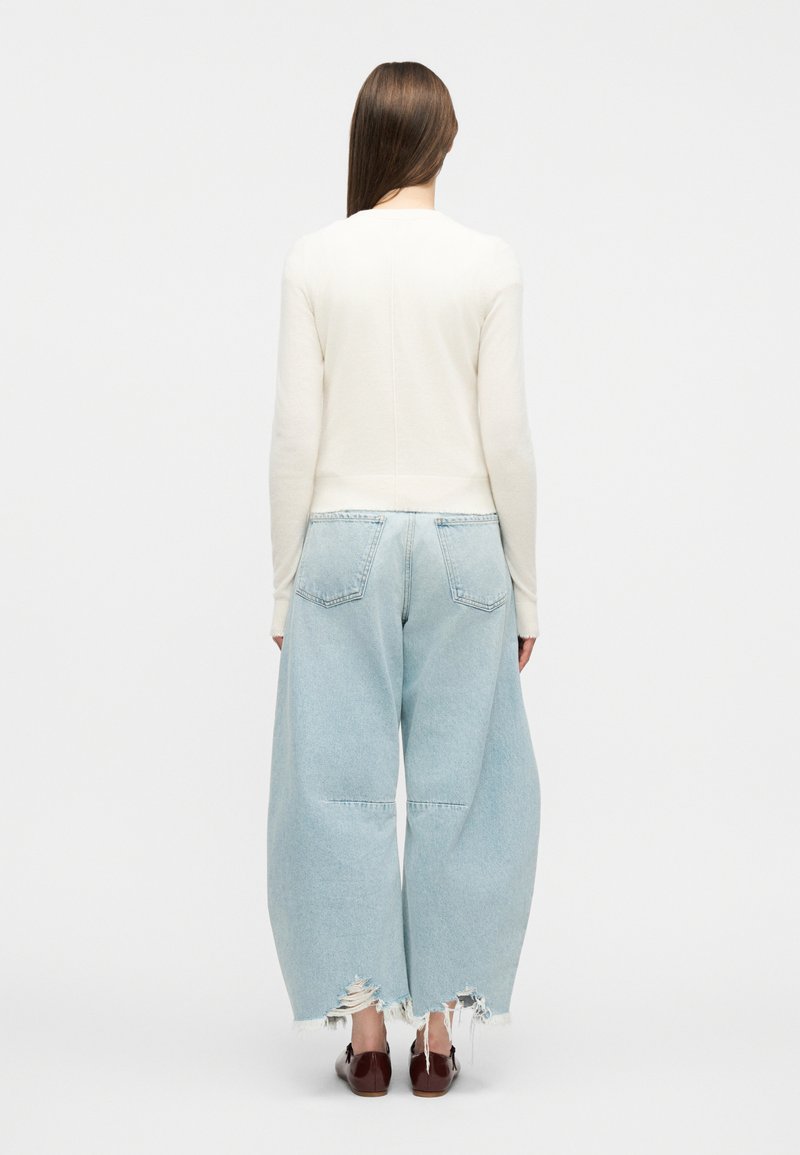 Person mit langem braunem Haar trägt einen weißen Pullover, weite, hellblaue, abgenutzte Jeans und braune Schuhe und steht vor einem einfarbigen Hintergrund.