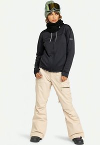 Sieviete, tērpta melnā aizdara jakā, bēšās sniega biksēs, pelēkā adītā cepurē, melnā šallē un slēpošanas brillēs, stāv ar rokām kabatās.