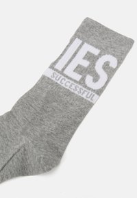 Szürke pamut zokni fehér felirattal, amelyen a „SUCCESSFUL” (Sikeres) szó olvasható, fölötte pedig egy sáv, amelyen a „LIES” (Hazugságok) szó található. Simára sodort felület.