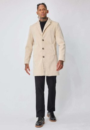 Manteau en laine beige avec col cranté, trois boutons, poches latérales et silhouette droite. Associé à un pantalon noir et des chaussures noires.