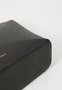 Black textured leather bag with a smooth surface, featuring an angled seam and gold logo detailing. Compact rectangular shape.