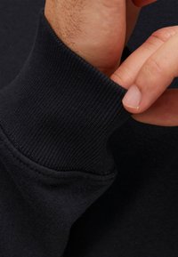 Polsino di felpa nera con texture a coste. Primo piano di una mano che regola la manica, mettendo in evidenza il tessuto morbido e i dettagli delle cuciture.