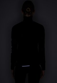 Svart jacka med hög krage och reflekterande detaljer på ärmar och nedre rygg, med en figurnära design, slät textur och långa ärmar.