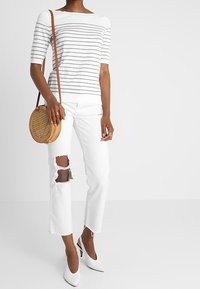Proužkovaný bílý a šedý pletený top, bílé džíny s roztrženými koleny, bílé podpatky a kulatá tkaná kabelka přes rameno.