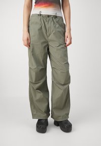 Oliwkowe, lekkie spodnie z szerokimi nogawkami, posiadające elastyczny pasek w talii i widoczne przeszycia; zestawione z czarnymi, perforowanymi klapami.