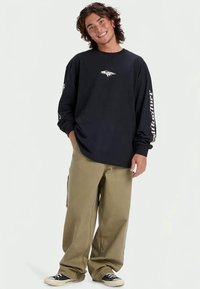 Czarna koszulka z długim rękawem z białym logo na klatce piersiowej i napisem "Quiksilver" na rękawach, w komplecie z luźnymi khaki spodniami i czarnymi trampkami.