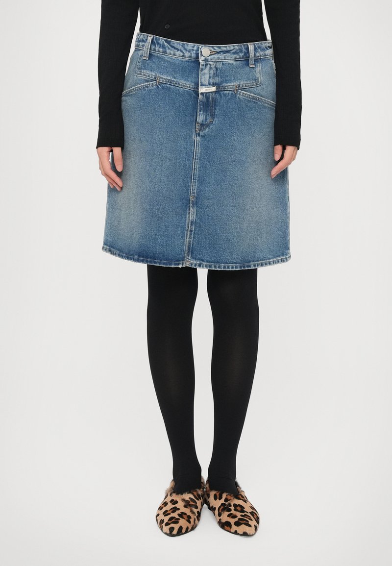 Denim skirt in blue, A-line shape, mid-thigh length, featuring two front pockets. Worn with black tights and leopard print slippers.