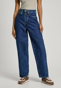 Jeans de ganga azul de cintura alta com um corte largo, costura em contraste e bolsos frontais, combinados com ténis castanhos e creme.