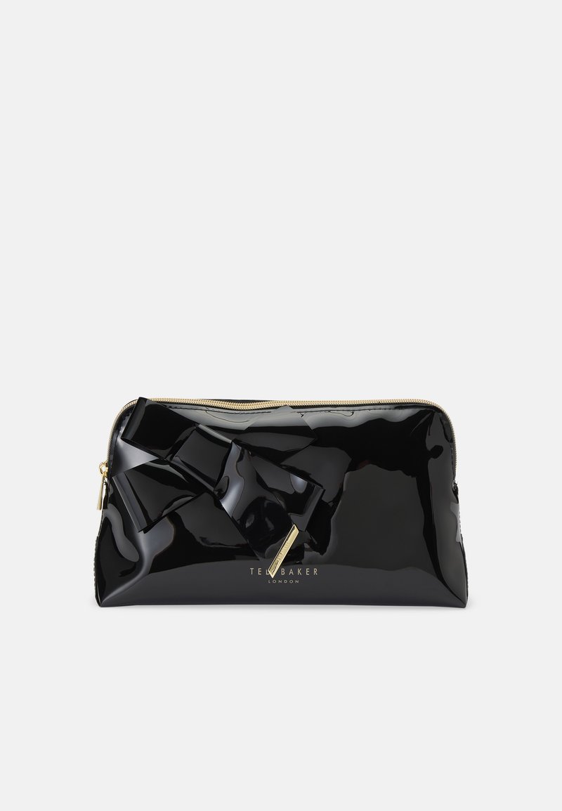 Bolsa de patentes pretas com um grande laço, detalhes em dourado e fecho com zíper. Apresenta um logótipo da marca em dourado na parte da frente.