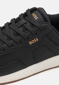 Svart sneaker med texturerat gummi och syntetmaterial, utrustad med platta svarta snören, guldfärgad "BOSS"-logotyp och en vit mönstrad sula.