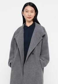 Grijze wolmix jas met een gelafd kraag, dubbele knopen en zijzakken, gedragen over een marineblauw overhemd met knopen. Glad textuur.