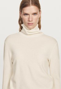Kremowy sweter z golfem wykonany z miękkiej dzianiny, z dopasowanym, złożonym kołnierzem oraz luźnym krojem i długimi rękawami.