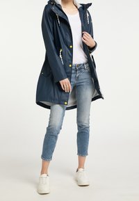 Marineblå vattentät regnkappa med huva, framfickor, kontrasterande gula knappar och justerbara dragsko; ihop med ljusblå jeans och vita sneakers.