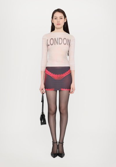 Durchsichtige Langarmbluse mit "LONDON"-Druck, kombiniert mit einem taillierten Rock mit rosa Spitzenakzenten, dazu schwarze Strumpfhosen und Absatzschuhe.