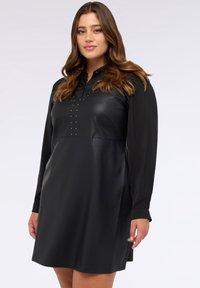 Schwarzes Kleid mit einem gerafften Leder-Oberteil, langen Ärmeln aus einem weichen Stoff und silbernen Nieten entlang der Vorderseite für zusätzliche Akzente.