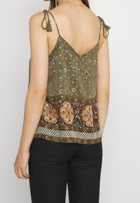 Grön blommönstrad linne med justerbara band och pom-pommar, med en dekorativ, mönstrad fåll med livliga färger och former.