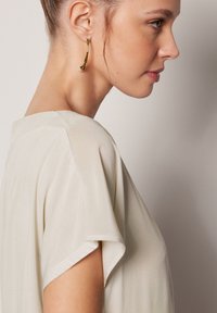 Ljusbeige topp med korta, draperade ärmar. Slät textur och avslappnad passform framhävd. Guldörhängen med en liten pärla som accent.