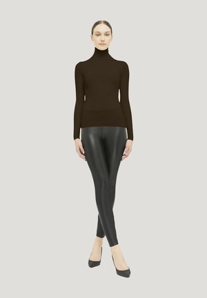 Vrouw staat met gekruiste benen, draagt een zwarte coltrui, zwarte leren leggings en zwarte pumps met puntige neus tegen een effen achtergrond.