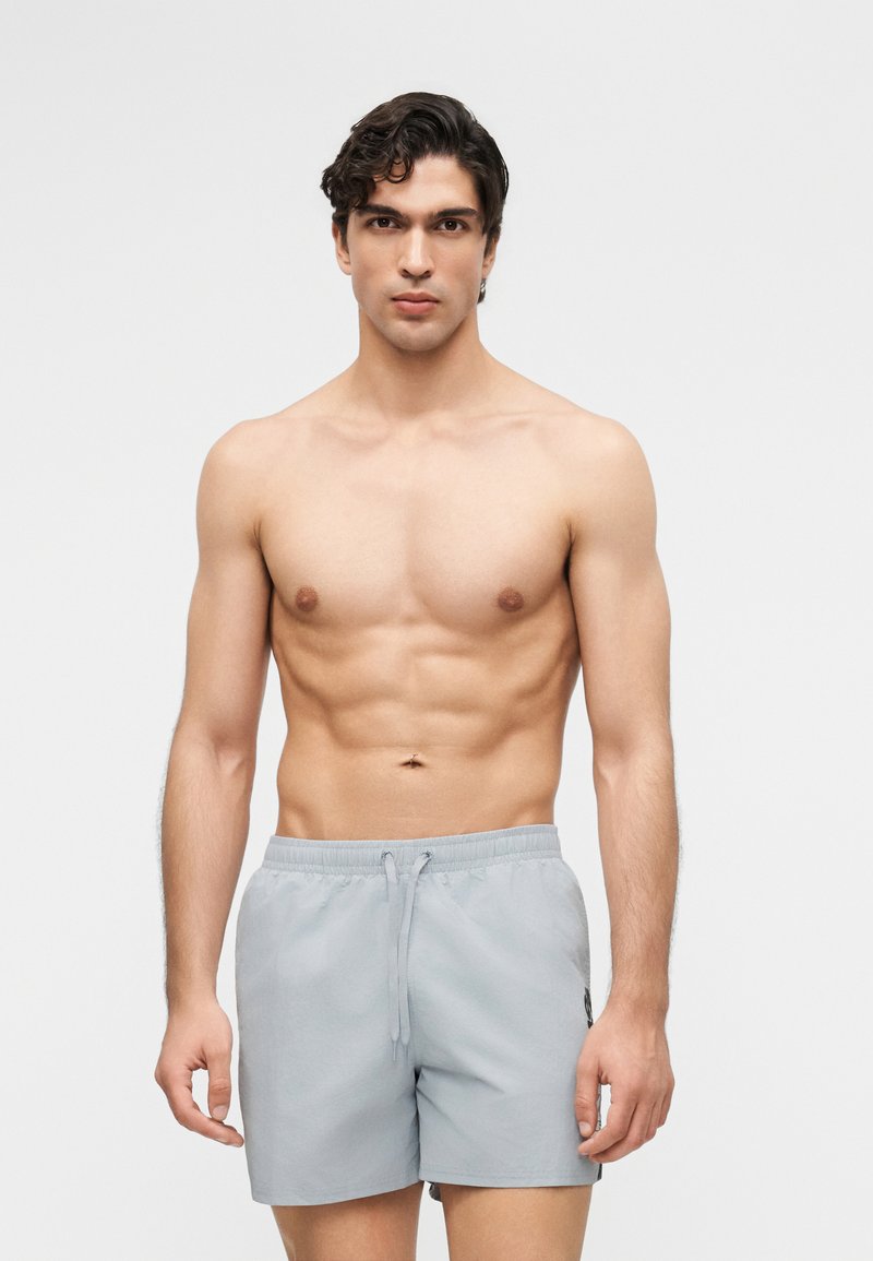 Αθλητικός νεαρός άνδρας με σκούρα μαλλιά, που φοράει ανοιχτό γκρι μαγιό, στέκεται μπροστά σε απλό λευκό φόντο.