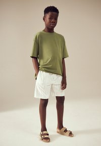 T-shirt en coton vert associé à un short blanc à fines rayures. Porte des sandales beiges avec deux sangles ajustables. Tenue d'été simple et décontractée.