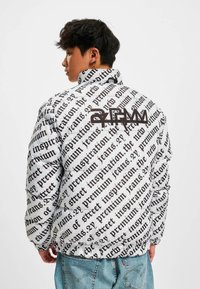 Jachetă din puf albă cu model de text negru, guler înalt și brand pronunțat pe spate. Textul are o tipografie îndrăzneață, mare.