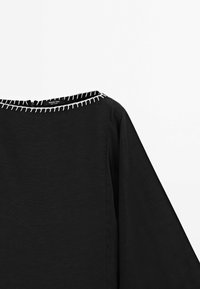 Blouse noire avec un large décolleté orné de surpiqûres blanches contrastantes. Design fluide, tissu léger et manches longues chauve-souris.