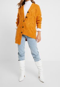 Cardigan arancione lavorato a maglia con un motivo a foglia testurizzato, caratterizzato da bottoni neri, indossato sopra una maglietta bianca e jeans blu, completato da stivali bianchi.