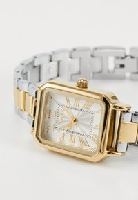 Hodinky ve tvaru obdélníku v zlaté a stříbrné barvě s vzorovaným stříbrným ciferníkem, římskými číslicemi a texturovanou zlatou korunkou na spojeném kovovém náramku.