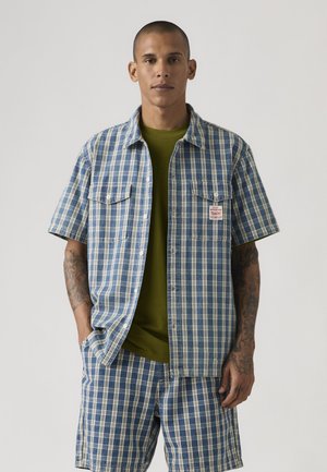 Man with tattoos wearing blue and white plaid short-sleeve shirt and matching shorts over olive green t-shirt, standing against plain background.