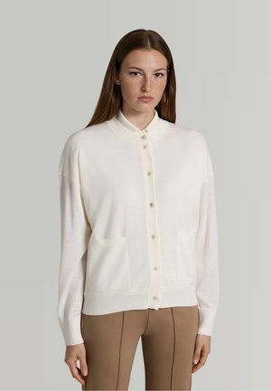 Jeune femme aux cheveux bruns lisses porte un cardigan blanc boutonné et un pantalon beige, debout devant un fond gris clair uni.