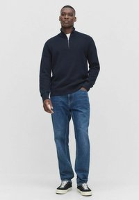 Morski sweter z zamkiem na szyi w teksturowanym dziewiarskim materiale, w połączeniu z niebieskimi dżinsami i czarnymi sneakersami. Stylizacja charakteryzuje się luźnym krojem i swobodnym stylem.