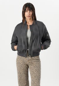 Chaqueta bomber negra con una textura suave y brillante. Presenta puños acanalados, un cierre frontal con cremallera y un bolsillo lateral. Llevada sobre una camiseta gris y pantalones de estampado de leopardo.