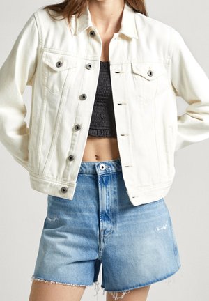 Kvinde iført en åben hvid denimjakke over en sort cropped top kombineret med højtsiddende blå denimshorts med frynsede kanter.