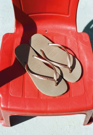 Beige Havaianas Flip-Flops mit strukturierten Sohlen, die auf einem roten Plastikstuhl liegen und Schatten auf eine helle Oberfläche werfen.