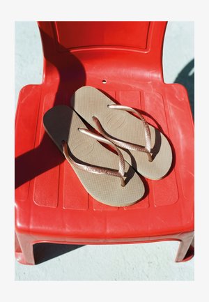 Beige Havaianas Flip-Flops mit strukturierten Sohlen, die auf einem roten Plastikstuhl liegen und Schatten auf eine helle Oberfläche werfen.