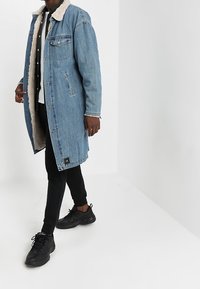 Abrigo largo de mezclilla con cuello de piel sintética beige, color azul, botones frontales y bolsillo en el pecho, combinado con pantalones negros y zapatillas.