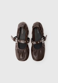 Ballerines en cuir marron foncé avec bords élastiques et brides à boucle réglables sur le dessus, vues de dessus sur un fond clair.