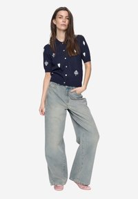 Námořnický krátký kardigan s bílými vzory srdcí a listů, zapínání na knoflíky, doplněný světle modrými širokými džínami a růžovými botami.