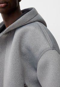 Grijze hoodie van een zachte, textuurrijke stof. Heeft een ruime capuchon en zichtbare stikseldetails langs de schouders. Ritsaccent aan de zijkant.