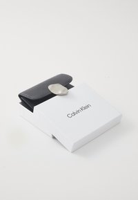 Musta nahast rahakott tekstureeritud viimistlusega ja hõbedane metallist lukuga, esitatud valges brändinguga karbis, millel on trükitud "Calvin Klein".