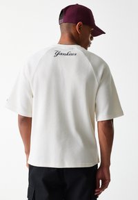 White, ribbed short-sleeve shirt featuring "Yankees" embroidered in cursive on the back; worn with a dark-colored cap.