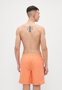 Young man with short dark hair, shirtless, wearing orange swim shorts, showing a fleur-de-lis tattoo on upper back, facing a plain white background.