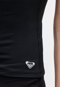 Μαύρο εφαρμοστό τοπ με μικρό λευκό λογότυπο σε σχήμα καρδιάς κοντά στο κάτω τελείωμα στην αριστερή πλευρά, φορεμένο από άτομο.