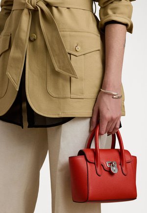 Sac à main en cuir rouge de forme arrondie, avec des accessoires en argent et des anses supérieures. Porté avec une veste beige et un pantalon couleur crème.