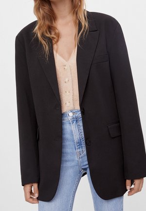 Woman wearing a black oversized blazer over a beige buttoned cardigan and light blue high-waisted jeans against a plain background.