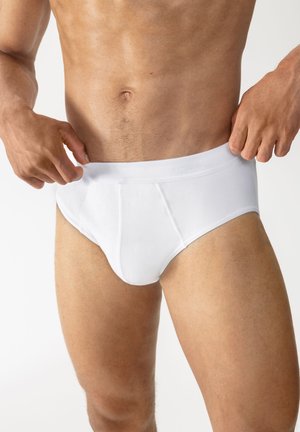 Mann in weißen Slips, der den Bund anpasst, zeigt einen durchtrainierten Oberkörper und obere Beine vor einfarbigem Hintergrund.