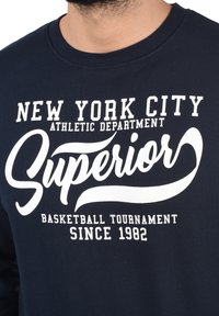 Mørkeblå sweatshirt med rund hals, prydet med hvid trykt tekst: "NEW YORK CITY ATHLETIC DEPARTMENT SUPERIOR BASKETBALL TOURNAMENT SINCE 1982."