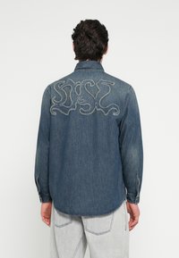 Πουκάμισο από τζιν σε σκούρο μπλε με μακριά μανίκια, με μεγάλο κεντητό σχέδιο στην πλάτη. Απαλή υφή, ίσια ραφή στο τελείωμα, μανσέτες με κουμπιά.